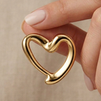 Gold heart-shaped earring held between fingers against a beige background
