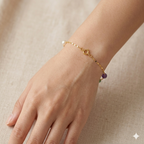 Gold bracelet with a purple bead on a hand against a beige background